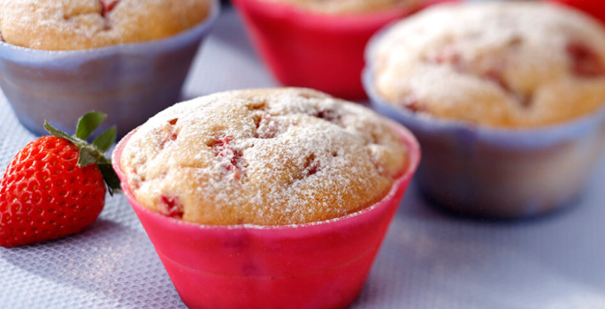 Fotografia em tons de vermelho e lilás em uma bancada com uma toalha emborrachada lilás, com forminhas pequenas vermelhas e lilás com os muffins de fubá com morango dentro delas.