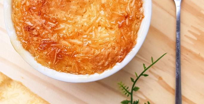 Foto da receita de Cocada de Forno. Observa-se um recipiente redondo branco com a cocada dentro assada e bem douradinha, sobre uma tábua de madeira