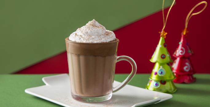 Fotografia em tons de marrom em uma bancada de madeira de cor verde. Ao centro, um bandeja branca contendo a bebida. Ao fundo, há 3 kikat sabor Natal.