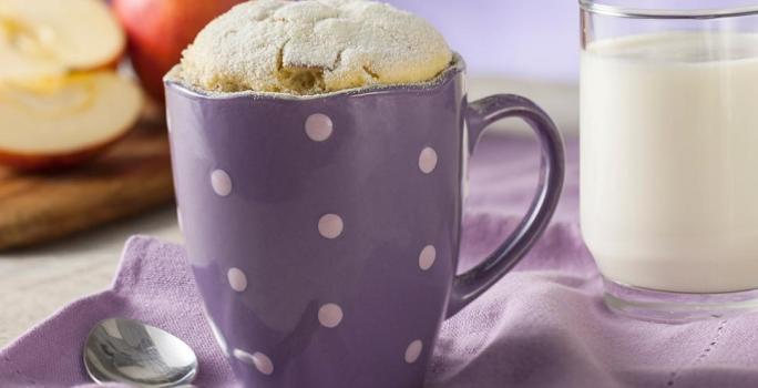 Fotografia em tons de roxo em uma bancada de madeira e um paninho roxo. Ao centro, uma caneca roxa de bolinhas brancas com o bolinho de caneca de leite Ninho em Pó. Ao fundo, maçãs cortadas ao meio e um copo de vidro com leite.