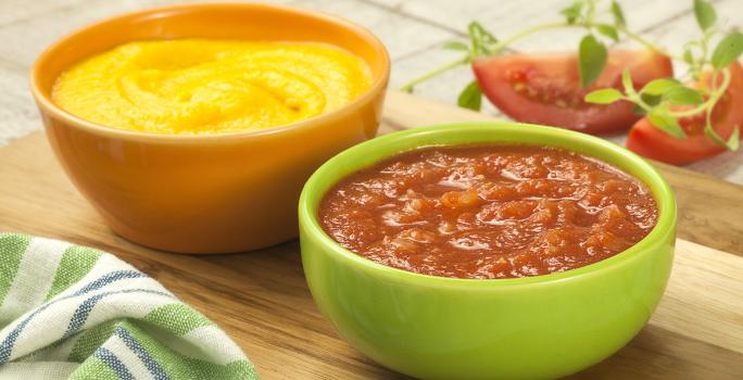 Fotografia em tons de verde, amarelo e vermelho com dois potes ao centro, sendo um amarelo com creme amarelo e um verde com um creme de tomate, ambos sobre peça de madeira, ao lado guardanapo e fatias de tomate, tudo sobre bancada de madeira.