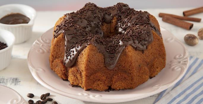 Foto da receita de Bolo de Cenoura com Especiarias Coberto com Brigadeiro de Café. Observa-se um bolo inteiro em cima de um prato de porcelana branco com brigadeiro por cima e granulado. Grãos de café, canela em pau e o brigadeiro decoram a foto.