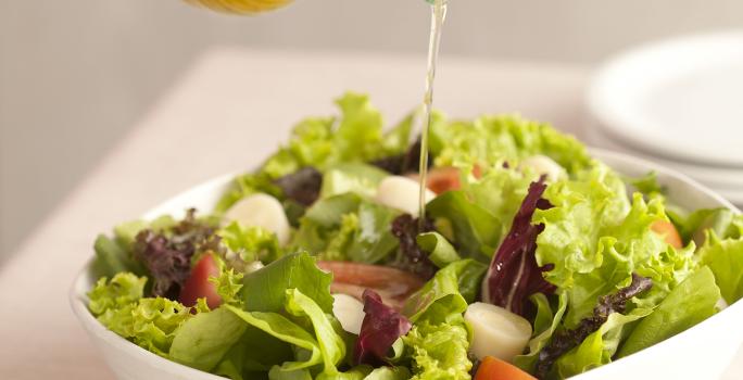 Fotografia em tons de verde e branco de uma bancada de madeira branca com um  recipiente branco quadrado com salada de folhas e um fio de azeite sendo despejado. Ao lado uma tabua de madeira com uma colher de servir.