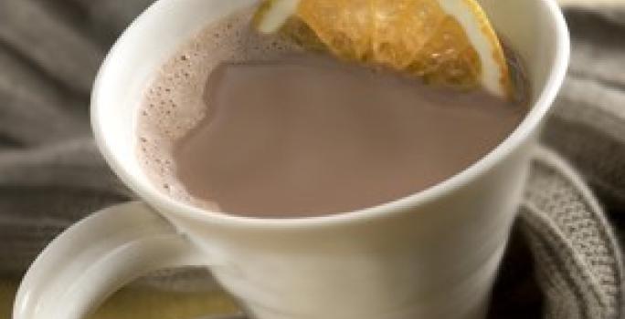 Fotografia em tons de cinza em uma bancada de madeira clara, um pano cinza escuro, uma xícara branca com o chocolate quente com uma rodela de laranja dentro.