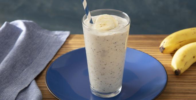Foto da receita de Smoothie de Banana com Farinha Láctea Nestlé. Observa-se um copo com a bebida e um canudo azul e branco. Ao lado direito, um cacho de bananas