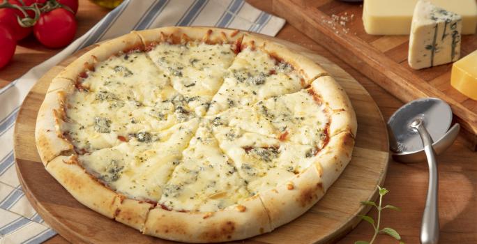Fotografia em tons de azul em uma bancada de madeira, um pano azul listrado, uma tábua de madeira de pizza, um cortador de pizza ao lado e pedaços de queijo.