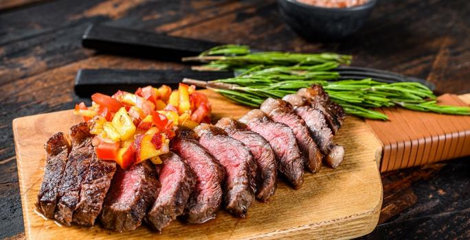 Fotografia em tons de marrom em uma bancada de madeira preta. Ao centro, uma tábua de madeira contendo a picanha fatiada ao fundo há duas facas, temperos e um recipiente contendo sal rosa.