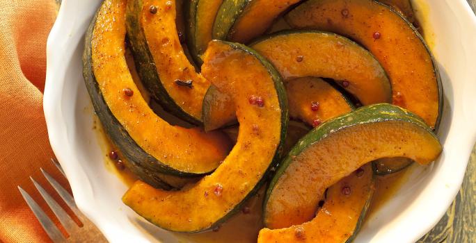 Fotografia em tons de laranja em uma bancada de madeira, ao lado um garfo apoiado em um guardanapo laranja e ao centro uma travessa redonda branca com as abóboras caramelizadas dentro.