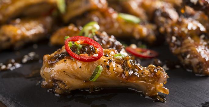 Fotografia de frango camponês dourado e brilhante com salsinha, gergelim e uma rodela de pimenta vermelha em cima, sobre um apoio preto. Ao fundo, mais unidades do frango.