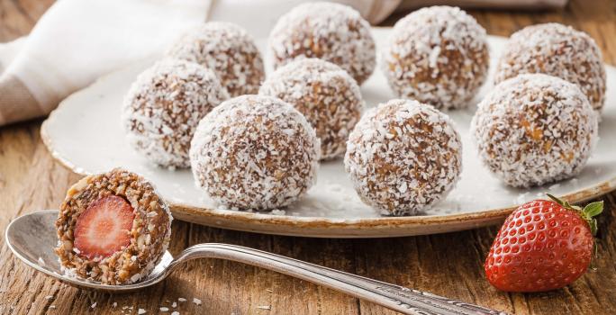 Fotografia em tons de branco, marrom e vermelho, ao centro prato branco com bombons enrolados em coco ralado, no entorno colher com bombom aberto ao meio, um morango, guardanapo branco com listras marrom, tudo sobre bancada de madeira rústica.