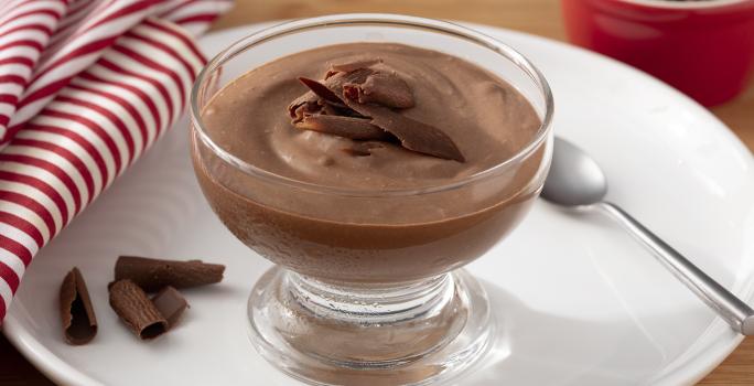 Fotografia em tons de marrom, branco e vermelho de uma bancada de madeira com um prato branco redondo, sobre ele uma taça com o mousse de chocolate zero, raspas de chocolate e uma colher. Ao lado um paninho listrado vermelho e branco.