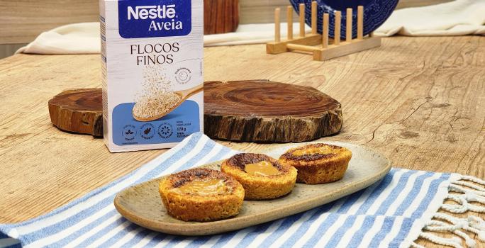 Fotografia em tons de marrom com um prato de cerâmica ao centro. Em cima do prato existe 3 muffins de doce de leite posicionados lado a lado. Ao fundo existe uma embalagem de Aveia Flocos Finos.