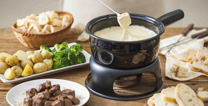 Fotografia em tons de marrom e beje de uma bancada de madeira com uma panela de fondue preta com fondue de queijo. Ao lado um prato redondo com carne, um prato retangular com batatas e brócolis, e uma cesta com pães.