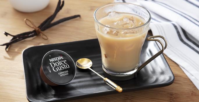 Foto em tons de bege da receita de espresso com baunilha e leite gelado servida em uma xícara de vidro sobre uma base de porcelana preta com uma colher dourada e uma cápsula de dolce gusto espresso intenso