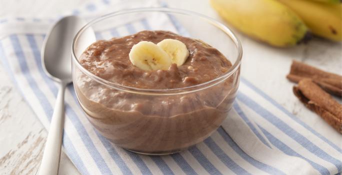 Fotografia em tons de azul em uma bancada de madeira clara com um paninho listrado azul e em cima um potinho de vidro com o doce de banana e cacau. Ao lado, algumas bananas, uma colher e canela em pau.