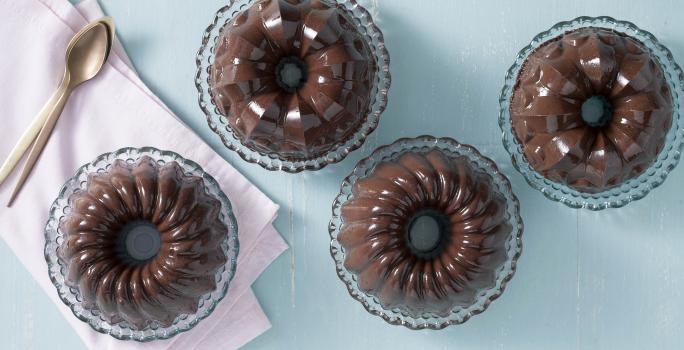 Fotografia em tons de chocolate, azul e rosa, com quatro porções de pudim de chocolate em pratinhos de cristal, um deles sobre guardanapo rosa tendo ao lado colheres de sobremesa, tudo sobre bancada azul.