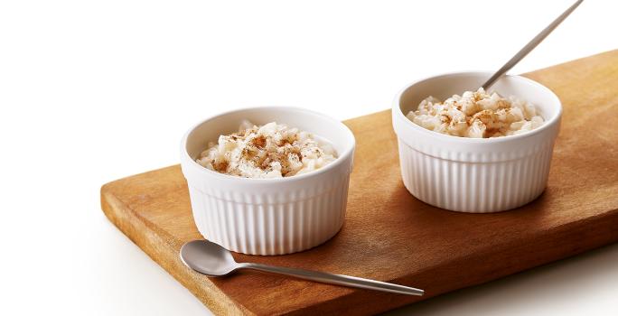 Foto da receita de Arroz Doce Vegano. Observa-se uma tábua de madeira com dois potinhos brancos e pequenos de arroz doce em cima.