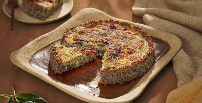 Foto da receita de quiche de aveia corte rústico servido em uma porção com uma fatia cortada sobre uma base quadrada de cerãmica marrom com um pano de linho bege ao lado