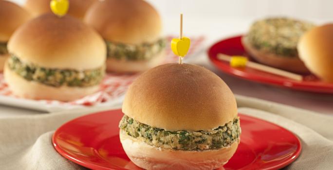 A foto contém diversos pães com hamburguês e palitos com uma bolinha amarela por cima. Um prato vermelho com um pão com hambúrguer à frente
