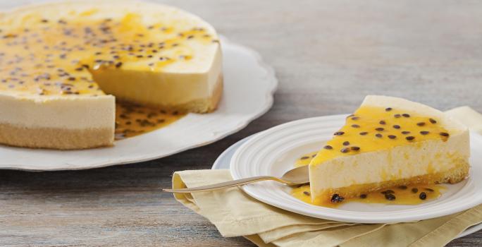 Fotografia em tons de amarelo e branco, com prato branco contendo uma torta de maracujá e ao lado um prato de sobremesa com uma porção da torta e uma colher de sobremesa, sobre guardanapo amarelo e outro prato, tudo sobre bancada cinza.