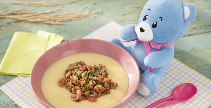 Foto da receita de Polenta Mole com Molho de Carne Moída. Observa-se um potinho rosa bebê com a polenta e a carne dentro, sobre um paninho quadriculado colorido. O ursinho Bo está do lado direito.