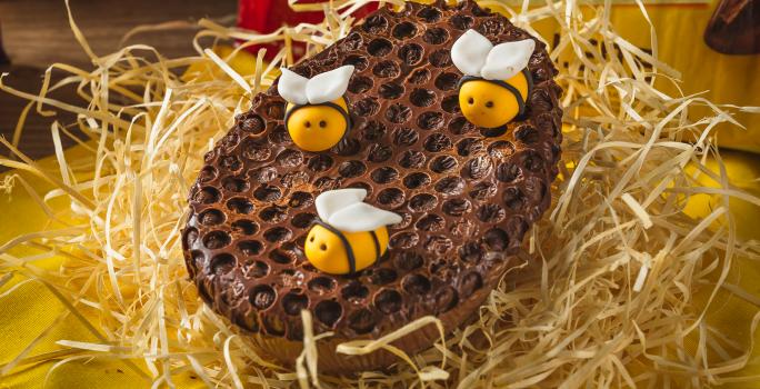 Foto aproximada de um ovo de chocolate de colher decorado com modelo de favo de mel e abelhas de mentira, sobre uma bancada com palha, tecido amarelo e uma barra de chocolate Garoto ao fundo