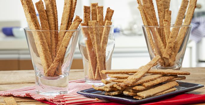 Fotografia em tons de pardo e vermelho de uma bancada de madeira marrom vista de frente, contém 3 copos transparentes e cada um contém biscoitos de cebolas em tiras. Ao lado uma travessa preta com mais biscoitos em tiras e abaixo um pano vermelho.