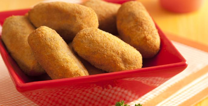 Fotografia em tons de amarelo e vermelho de uma bancada de madeira com um pano dobrado quadriculado branco e laranja, um recipiente vermelho quadrado com vários bolinhos fritos e ao fundo dois copos rosa.