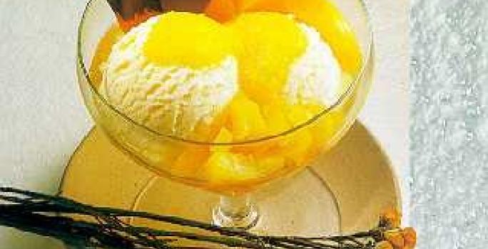 Fotografia em tons de amarelo em uma bancada de madeira, um pano bege, um prato redondo raso bege com uma taça de vidro com o sorvete de creme e pêssegos em calda dentro dela. Ao lado, flores amarelas e laranjas.