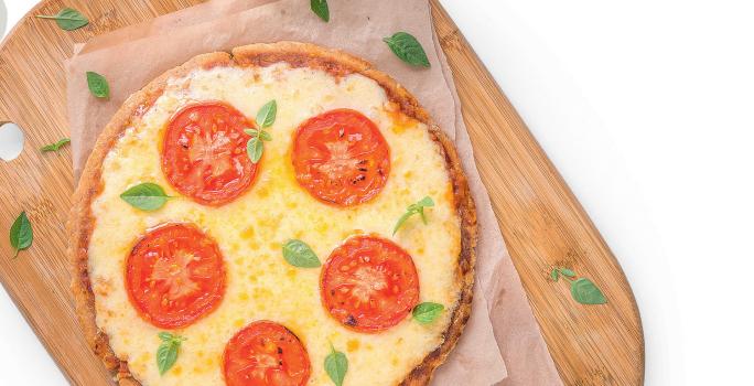 Fotografia em tons de amarelo e vermelho em uma mesa branca com uma tábua de madeira, um pano bege e uma pizza média de queijo com tomate em cima dela.