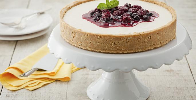 fotografia em tons de branco, bege e roxo de uma bancada cinza vista de frente, contém um suporte para servir sobremesas branco com a torta com calda de frutas vermelhas por cima, ao lado um pano laranja com uma espátula e 2 pratos ao fundo branco.