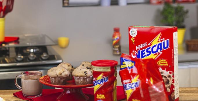 Foto vista de frente para um bancada com produtos Nescau à direita, mais ao centro um prato vermelho alta com quatro Muffins Crocantes de Nescau e à esquerda uma caneca com leite e Nescau.