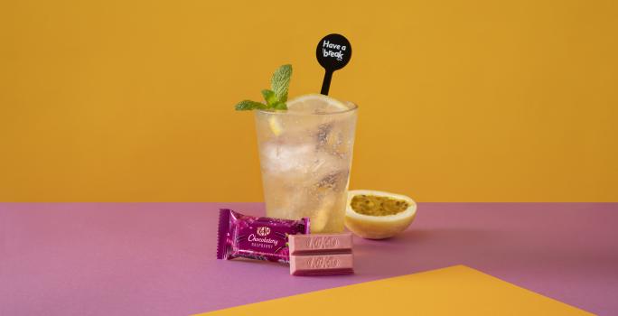 Foto da receita de Soda Italiana Passion Fruit and Lemon. Observa-se um fundo amarelo e roxo com um copo alto no centro, cheio de gelo, com uma rodela de limão siciliano. Meio maracujá decora a foto à direita, assim como folhas de hortelã no copo.