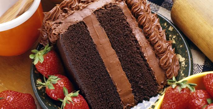 Fotografia de uma fatia de bolo com massa e recheio de chocolate, morangos por toda a parte em um prato desenhado de cor escura, apoiado em uma toalha de mesa azul e branca, quadriculada. Ao lado do bolo, dois potes e um rolo de massa.