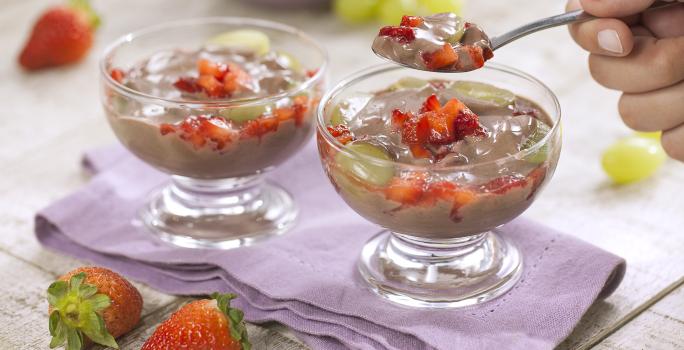 Fotografia em tons de branco e roxo de uma bancada de madeira branca com um paninho roxo, sobre ele duas taças de frutas com creme e uma colher retirando um pedaço. Ao fundo uvas e morangos. A frente dois morangos.