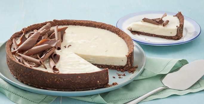 Fotografia em tons de azul em uma mesa de madeira azul com um prato raso azul claro e a torta de limão e chocolate. Ao lado, um prato pequeno com uma fatia da torta e uma espátula de bolo.