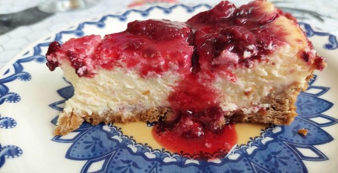 Foto em tons de vermelho da receita de cheesecake de frutas vermelhas ao estilo nova york servida em uma fatia sobre um prato decorado de porcelana azul e branco