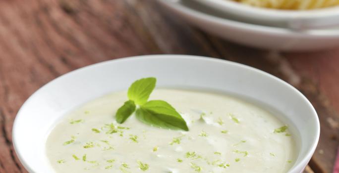 Fotografia com tons de branco e marrom, com ramequim branco com molho, ao fundo prato branco com macarrão parafuso e pedaço de queijo com ralador sobre tábua branca, ao lado guardanapo listrado, tudo sobre bancada de madeira rústica.,