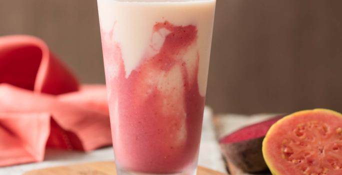 Fotografia em tons de rosa em uma bancada de madeira, um pano rosa, uma tábua de madeira, um copo de vidro alto com a bebida de iogurte com goiaba, morango e beterraba. Ao lado, uma goiaba cortada ao meio.