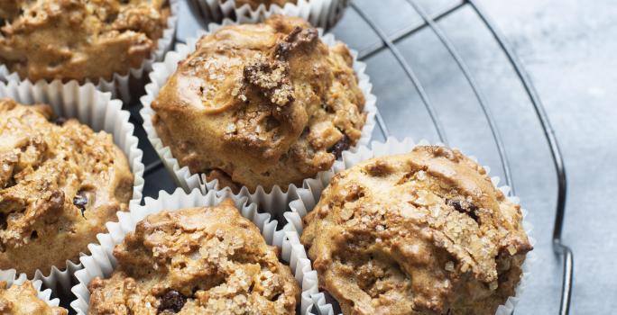 Fotografia de sete muffins com chocolate branco e preto em uma forminha de papel branca, que estão em cima de uma grade redonda na cor preta. Os bolinhos estão sobre uma mesa de cor clara.