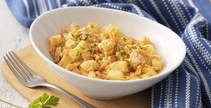 Fotografia em tons de azul em uma bancada de madeira com um pano listrado azul ao lado. No centro, um prato fundo branco com a salada de batata, atum, cebola e salsinha.