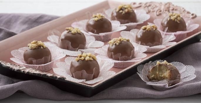 Fotografia em tons de lilás e chocolate, com bandeja lilás claro ao centro contendo bombons de chocolate sobre forminhas rendadas brancas e decorados com nozes picadas, ao lado bombom mordido, sobre guardanapo lilás, tudo bancada em tom lilás.