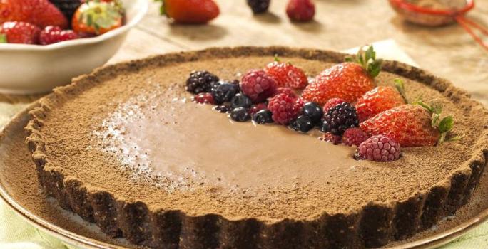 Fotografia em tons de marrom em uma mesa com uma toalha bege, um prato raso redondo grande com a torta de biscoito Nesfit com cacau e frutas vermelhas para decorar a torta, em cima do prato.