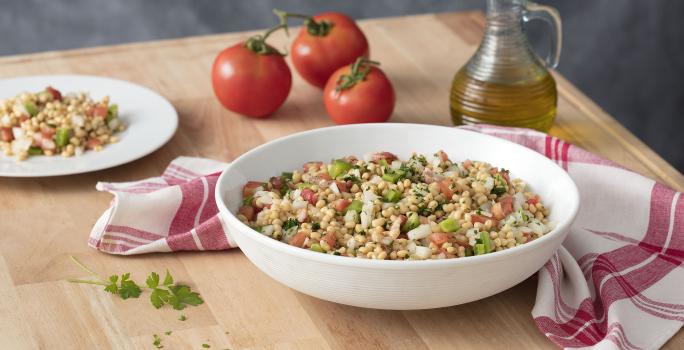 Fotografia em tons de branco e vermelho, ao centro recipiente branco com salada de feijão e tomate, ladeada por prato com salada, três tomates, galheteiro com azeite e guardanapo branco com listras vermelhas, tudo sobre bancada de madeira e fundo cinza.