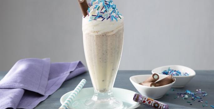 Foto da receita de milkshake algodão doce servida em um copo largo e alto de vidro sobre uma mesa acinzentada com um paninho lilás ao lado, além de algumas unidades de baton