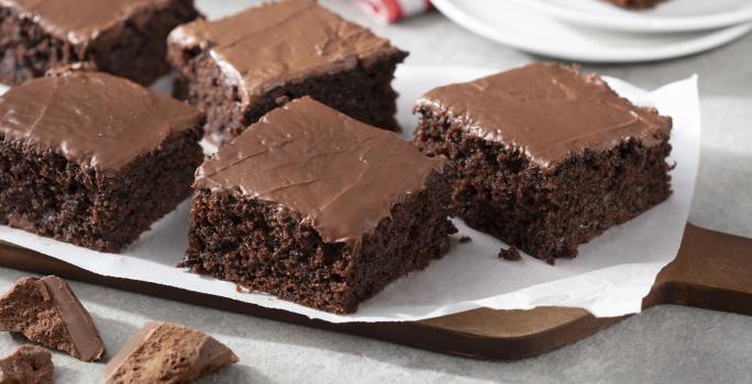 Fotografia em tons de marrom e branco de uma bancada branca  com uma tábua de madeira, sobre ela um papel manteiga com pedaços de bolo de chocolate zero. Ao fundo dois pratos brancos com uma fatia de bolo de chocolate e um paninho vermelho e branco.