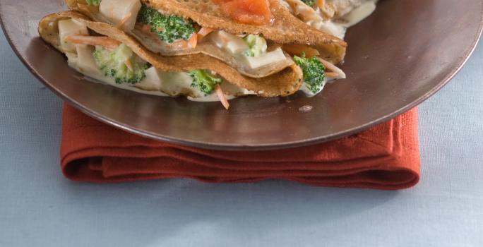 Fotografia de um prato marrom com 2 panquecas feitas em forma de crepe e recheadas com legumes e cobertas com molho de tomate, sobre guardanapo vermelho e toalha azul clara.