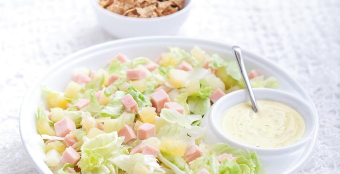 Fotografia em tons de branco e verde de uma bancada de madeira branca com paninho branco, sobre ele um recipiente redondo branco com a salada tropical e com o molho de iogurte. Ao fundo um recipiente redondo branco com o Nesfit Cereal.