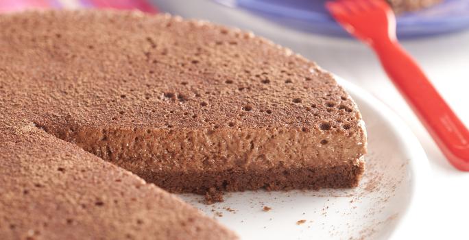 fotografia em tons de azul e marrom tirada de uma torta de chocolate com uma fatia retirada, e ao fundo um prato redondo azul com um pedaço da torta