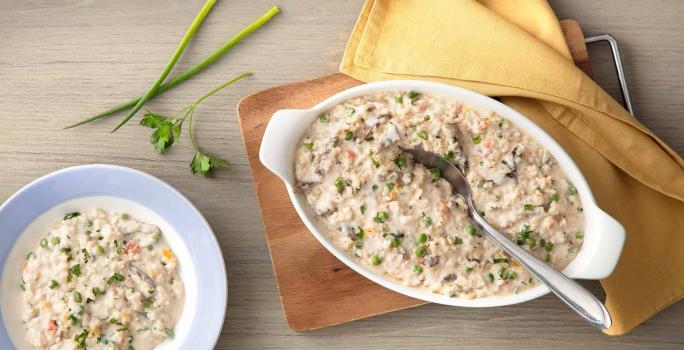 fotografia em tons de cinza e bege de uma bancada cinza vista de cima. Contém uma tábua de madeira com um recipiente oval branco com alças que contém o arroz  com creme, pedaços de carne e ervilhas por cima e ao lado um pano amarelo.